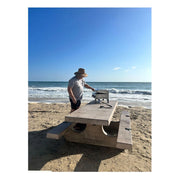 Kokomo Grills On The Go portable stainless steel grill with dual burners, carry handle, and locking clasps on a beach picnic table.