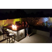 Kokomo Grills Baja/Fire L-Shaped BBQ Island with fire pit, 4 burner grill, outdoor fridge, and illuminated countertop at night.