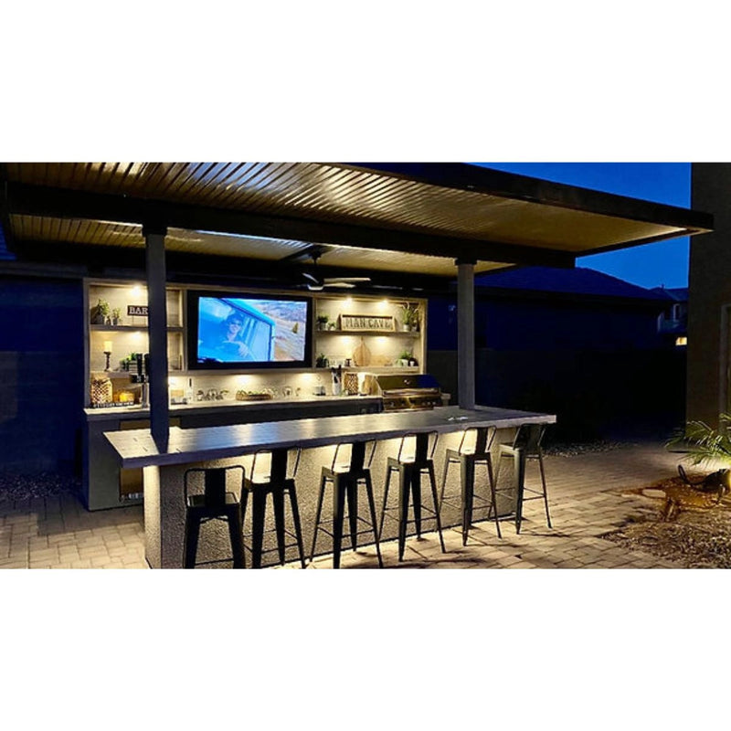 Kokomo Grills Fantasy Island 20'x20' outdoor kitchen with 5-burner grill, 70" TV, aluminum roof, bar seating, and bar lighting at night.
