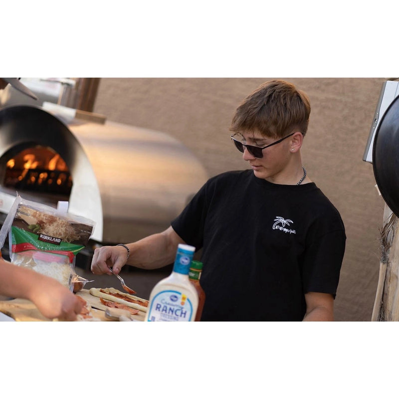 Kokomo Grills 32 Inch Dual Fuel Gas or Wood Fired Stainless Steel Pizza Oven with oven cart and built-in temperature gauge cooking outdoors.