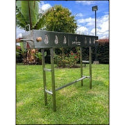Grill Brothers Robatta freestanding stainless steel charcoal grill with cutout designs and removable legs on a grassy lawn.