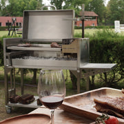 Tagwood BBQ Freestanding Argentine Santa Maria Grill in stainless steel with height-adjustable grates and gaucho-style brasero firebox outdoors.