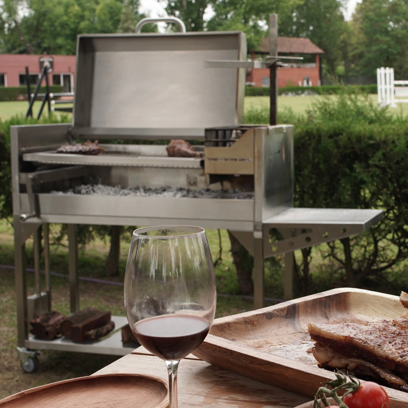 Tagwood BBQ Freestanding Argentine Santa Maria Grill in stainless steel with height-adjustable grates and gaucho-style brasero firebox outdoors.