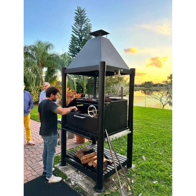 Tagwood BBQ The Beast Argentine Santa Maria Style black painted steel grill with height-adjustable grates and peaked roof outdoors.