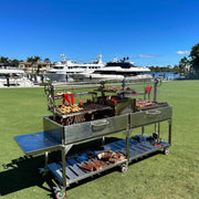 Tagwood BBQ XL Freestanding Argentine Santa Maria grill in stainless steel with double cooking space, height-adjustable grates, and wheels.