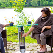Two people sitting outdoors by a table with a patio heater, surrounded by greenery.