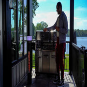 Person using a grill on a balcony with a scenic view of water and trees.
