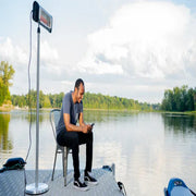Man sitting on a chair by a lake using a smartphone with a solar panel on a stand.
