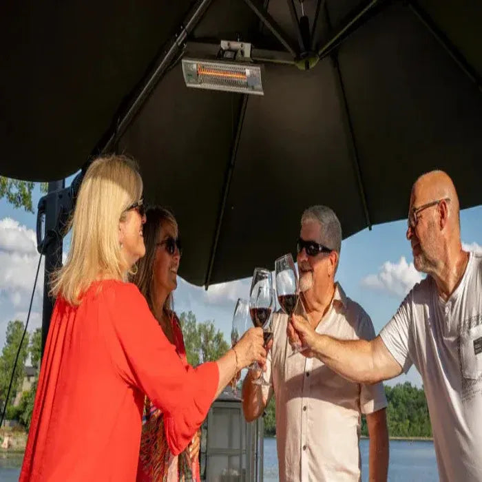 Group of people toasting with wine glasses under a black outdoor umbrella by a body of water.